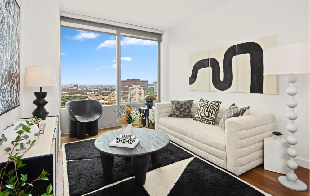 An apartment living room, furnished in black and white decor, with a view from a large window of the city below.