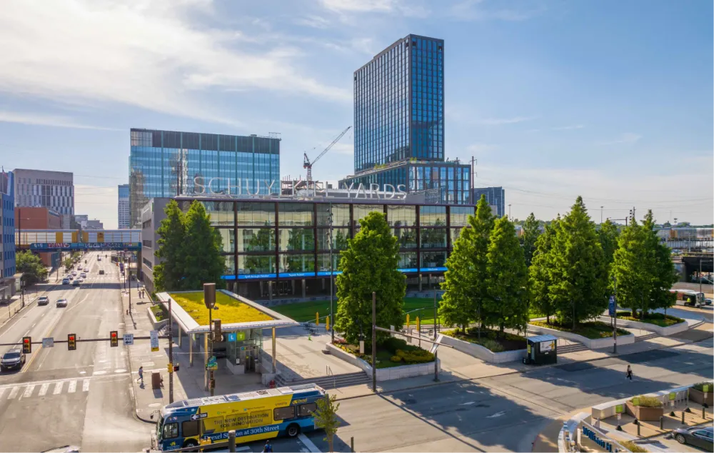 An exterior view of the Schuylkill Yards building showing the outdoor space including a covered seating area, planters with trees, and a grassy area.